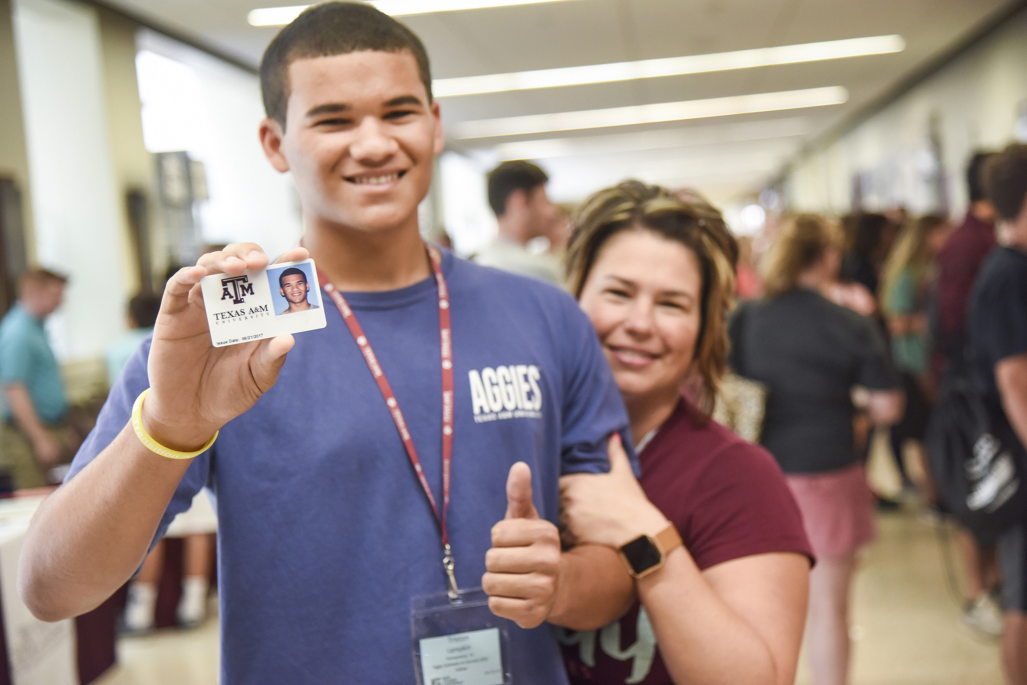 A male incoming student smiling and showing his Aggie card with his mom standing next to him.